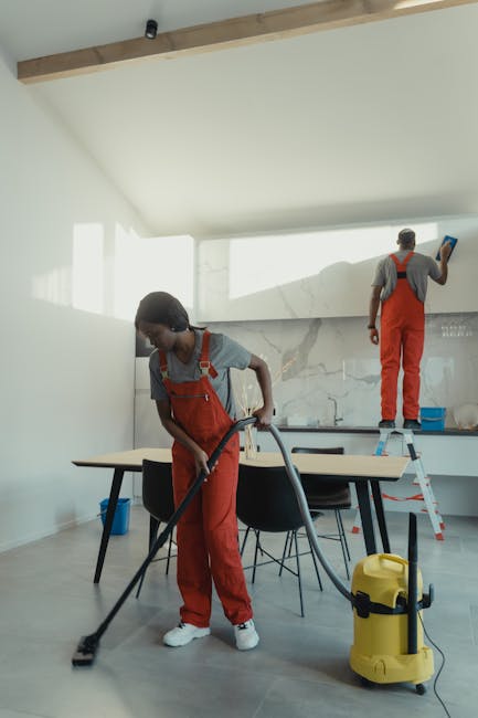 A person kneeling on a patterned area rug in a residential setting, preparing a yellow and black vacuum cleaner for use. The individual is wearing a beige jacket and blue jeans, and their hands are ad