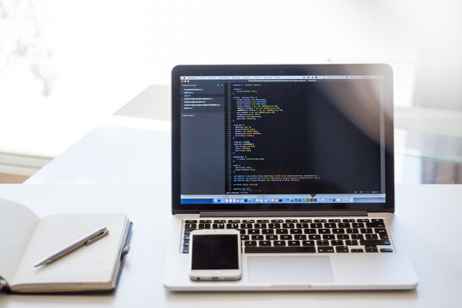 A silver laptop on a white desk displaying lines of programming code on a dark background, with a closed white notebook and a silver pen placed beside it, set in a well-lit room with natural sunlight 